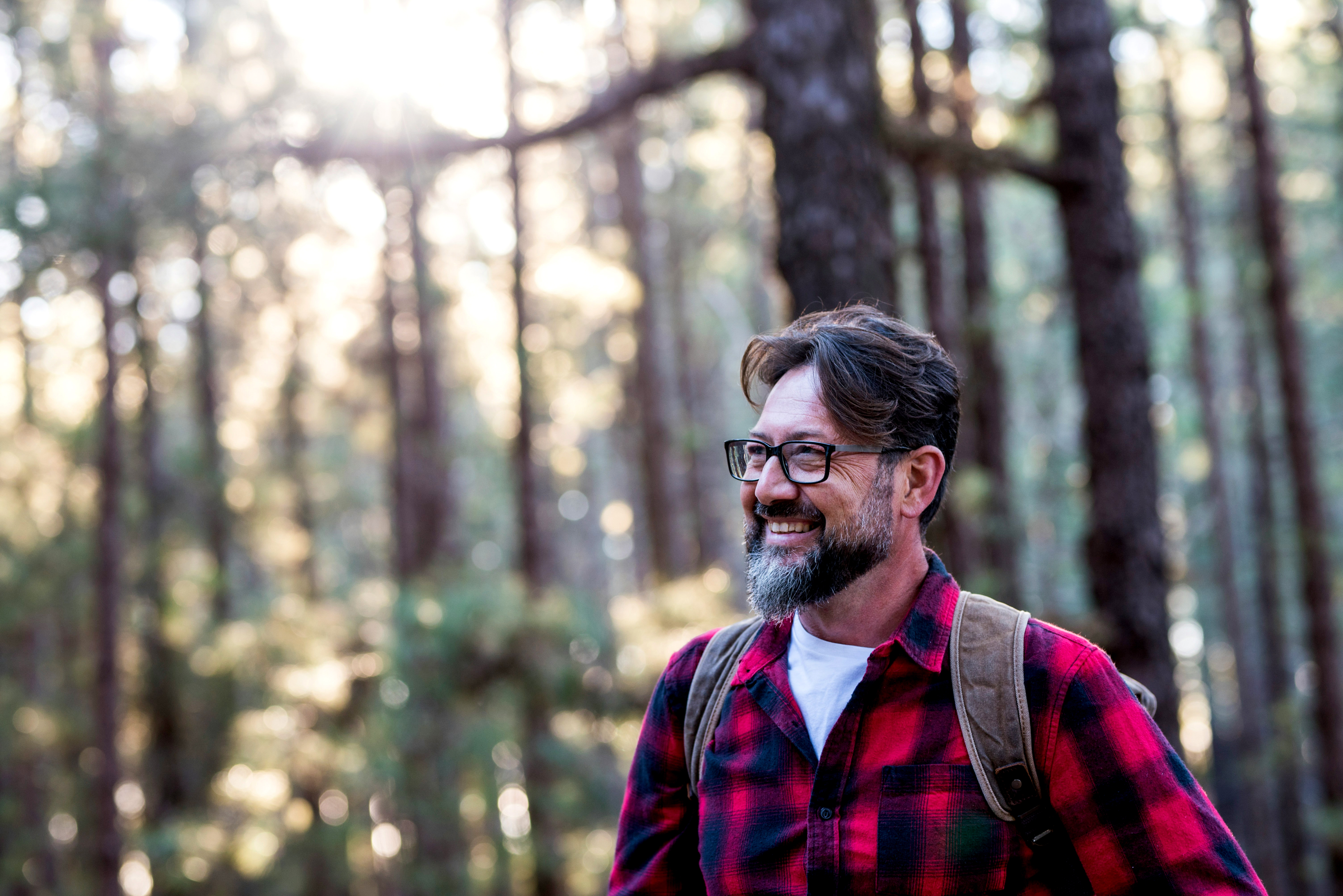 A middle aged man wearing a red checked flannel shirt and a backpack stands smiling in a forest
